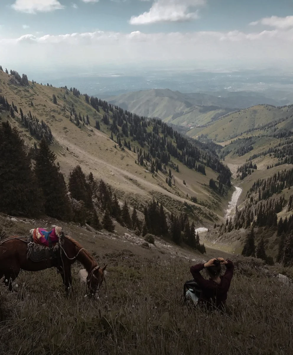 Traveler resting on a grassy slope beside a horse with panoramic view of Medeu Gorge and Almaty below
